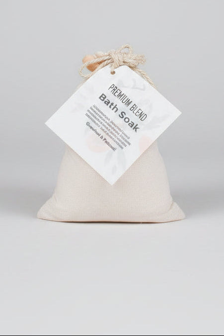 A small soft pouch of bath soak with a hanging paper tag reading 'Premium Blend BATH SOAK Grapefruit & peach,' tied with twine, on a wooden surface.